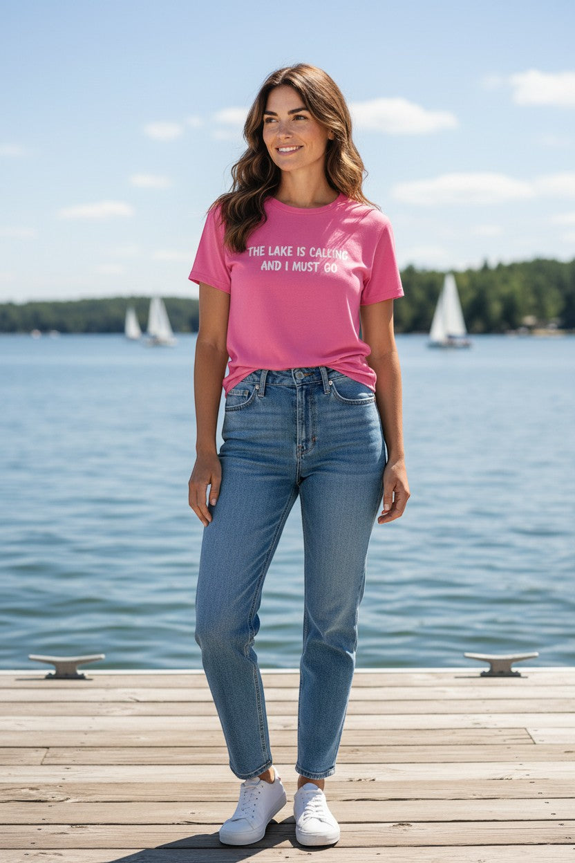 A woman wearing a pink Lakelifewear T-shirt with the text The Lake Is Calling and I Must Go on a wooden dock.