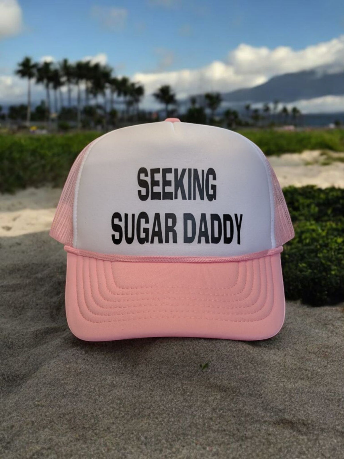 Lakelifewear white and pink foam trucker hat with Seeking Sugar Daddy slogan in black text on a sandy beach.